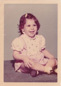 Faded image of a young female child sitting on a carpet. Her legs are crossed and her hands on her knees. She is wearing a pink dress. She has dark hair and a barette is holding back her bangs.