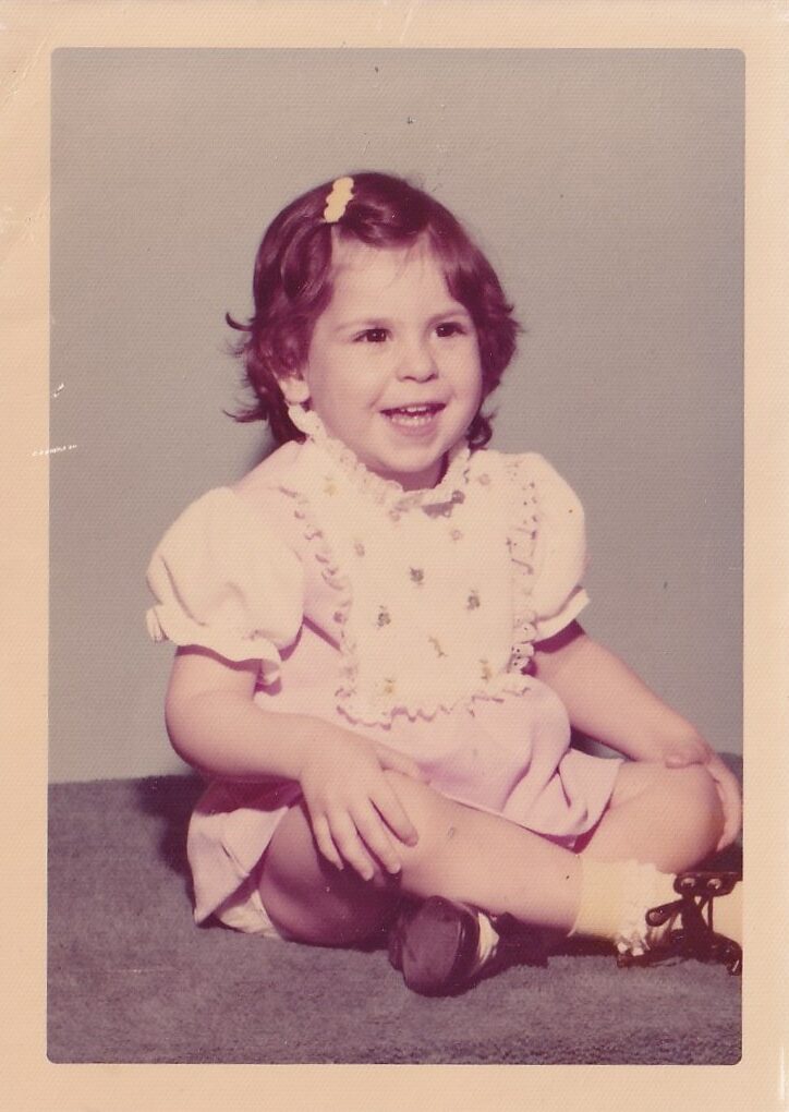 Faded image of a young female child sitting on a carpet. Her legs are crossed and her hands on her knees. She is wearing a pink dress. She has dark hair and a barette is holding back her bangs.
