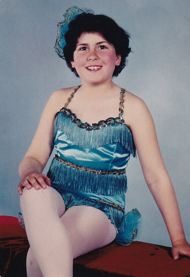Color image of a young girl with dark hair and dark eyes. She is sitting on a block with her legs crossed and her right hand on her right knee. She is wearing a sparkly blue dance outfit and has a sparkly blue bow in her hair.